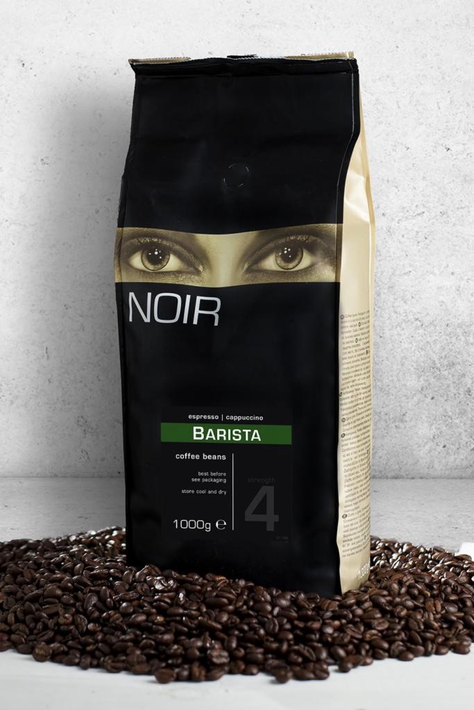 Czarne opakowanie kawy ziarnistej Noir Barista na tle ziarenek kawy i szarej ściany.