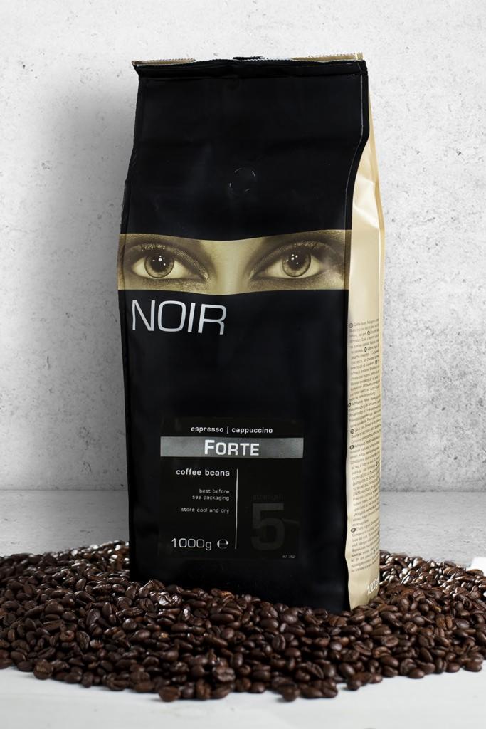 Czarna paczka kawy ziarnistej Noir Forte espresso/cappuccino, 1000g, na tle rozsypanych ziaren kawy i szarej ściany.