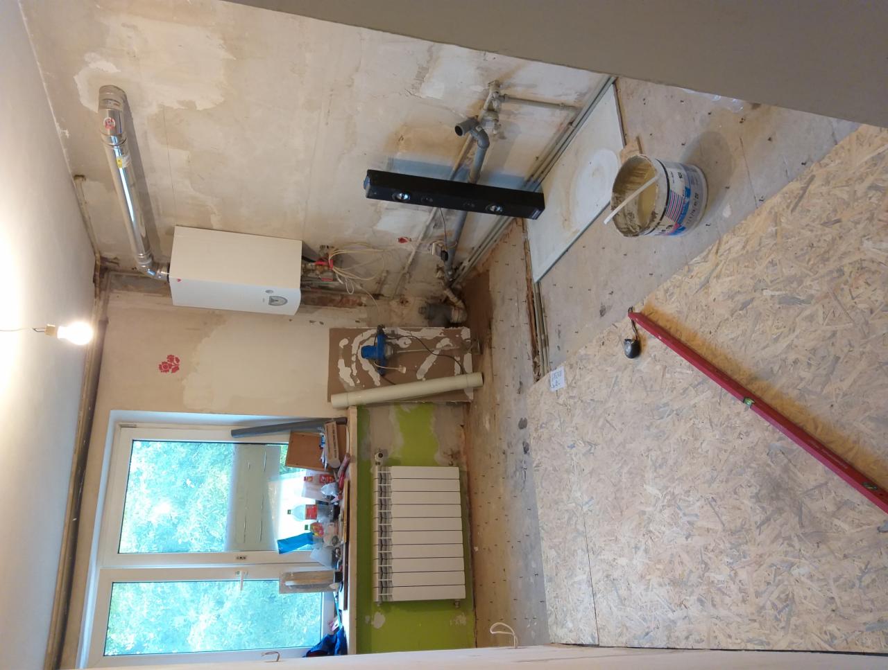 Wnętrze remontowanego pomieszczenia z widocznymi elementami instalacji, grzejnikiem, oknem, płytą OSB na podłodze, wiadrem z masą szpachlową i poziomicą.