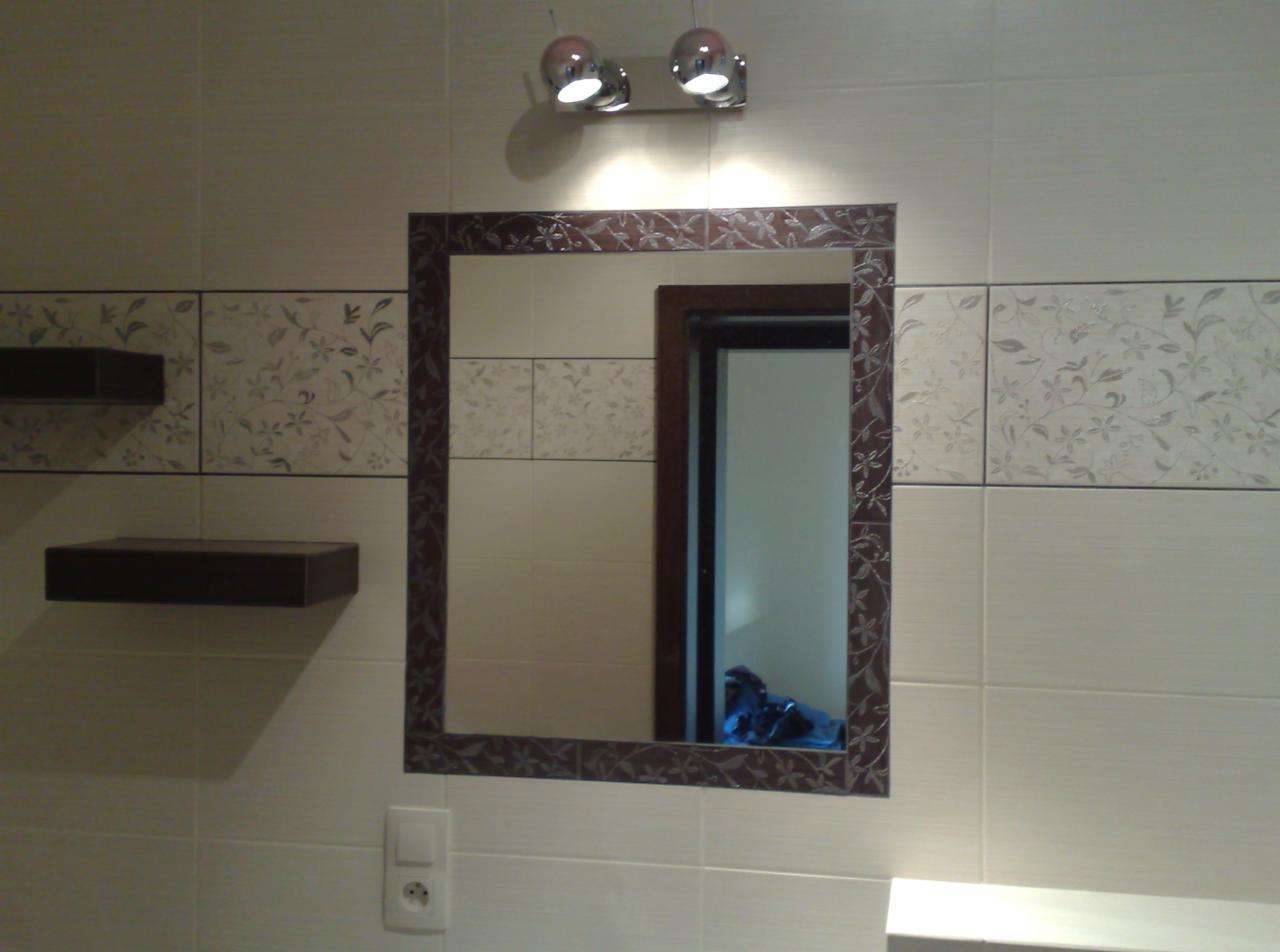 Ściana łazienki z beżowymi płytkami, dekoracyjnym pasem z motywem roślinnym, lustrem w brązowej ramie z motywem liści, oświetleniem punktowym i półkami wiszącymi.