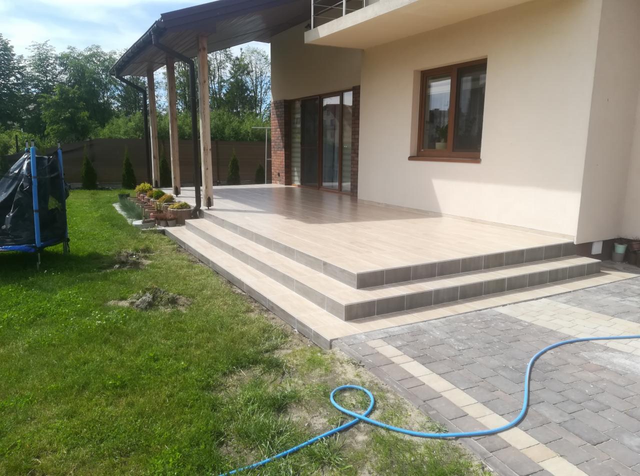 Wyłożony jasnymi płytkami taras z trzema stopniami, obok trawnik, błękitny wąż ogrodowy na kostce brukowej.