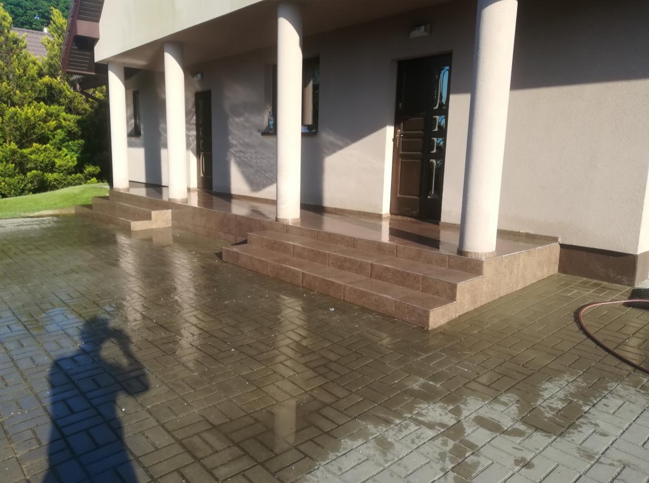 Wyłożone płytkami schody wejściowe do budynku z kolumnami, mokra kostka brukowa na podjeździe, cień osoby fotografującej w lewym dolnym rogu.