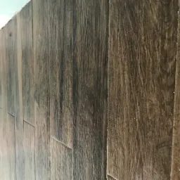 Ściana wyłożona panelami imitującymi ciemne, postarzane drewno, widoczne drobne niedoskonałości i szczeliny między panelami, fragment surowej ściany w tle.