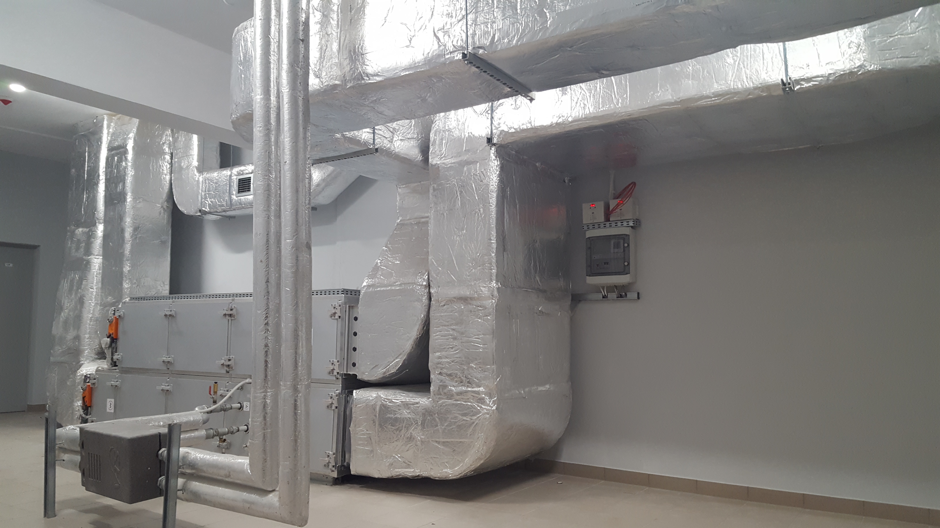 System kanałów wentylacyjnych obłożonych folią izolacyjną w pomieszczeniu technicznym, widoczna skrzynka elektryczna na ścianie.