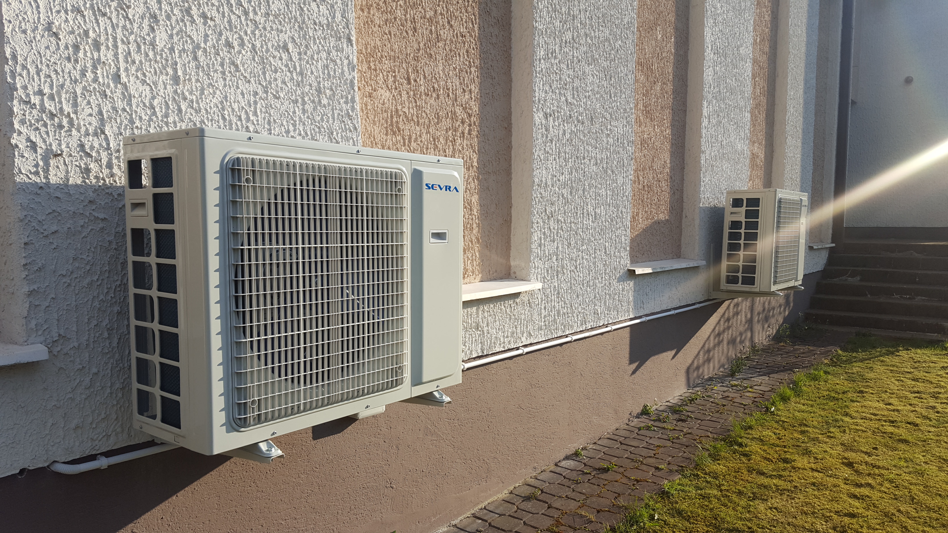 Dwa klimatyzatory zewnętrzne marki SEVRA zamontowane na elewacji budynku z ozdobnym tynkiem w pasy, widoczne przewody i kostka brukowa.