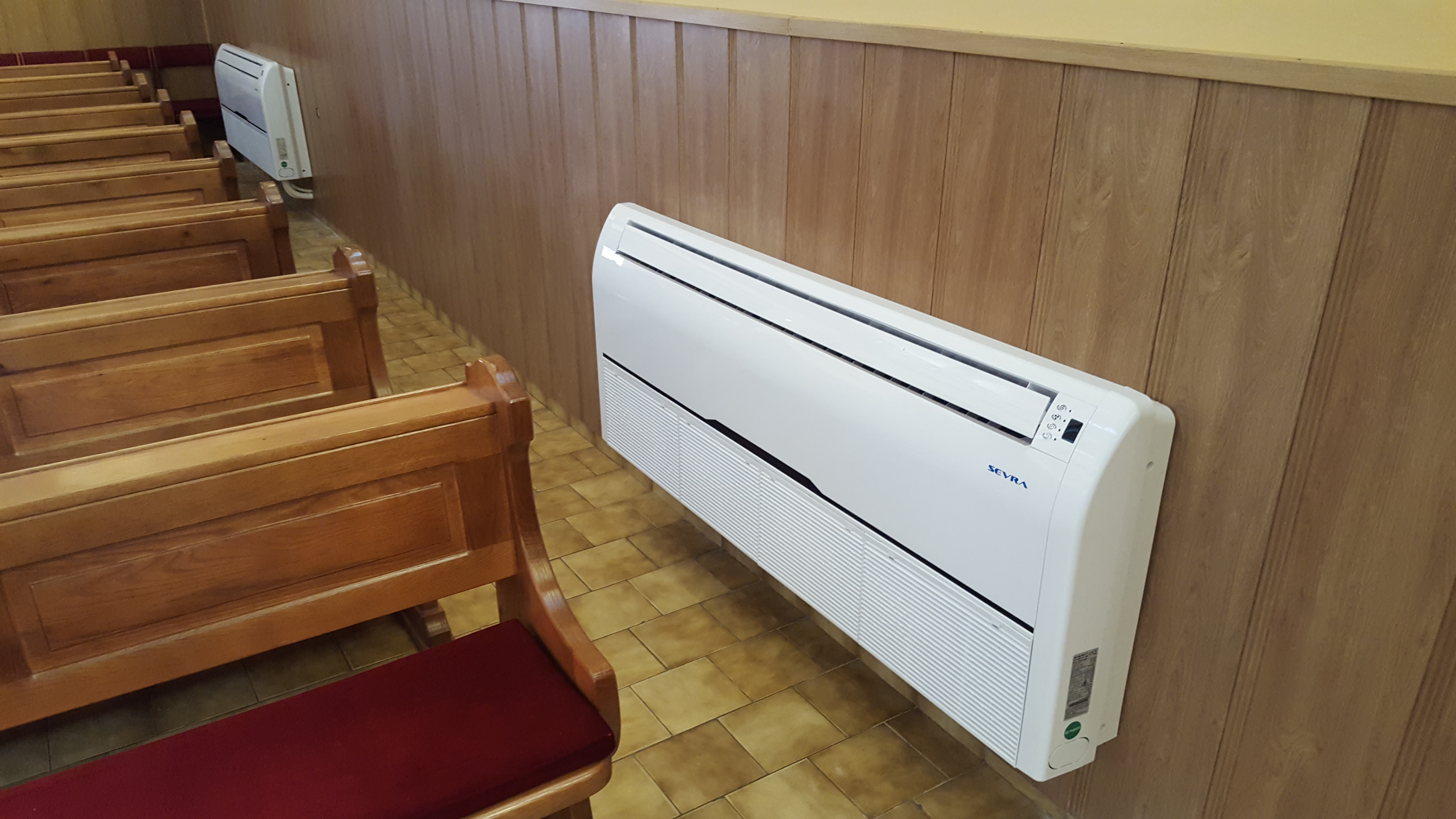 Biała jednostka klimatyzacyjna zamontowana na drewnianej ścianie w pomieszczeniu z ławkami. Widoczna marka SEVRA. Drugi, identyczny klimatyzator w tle.