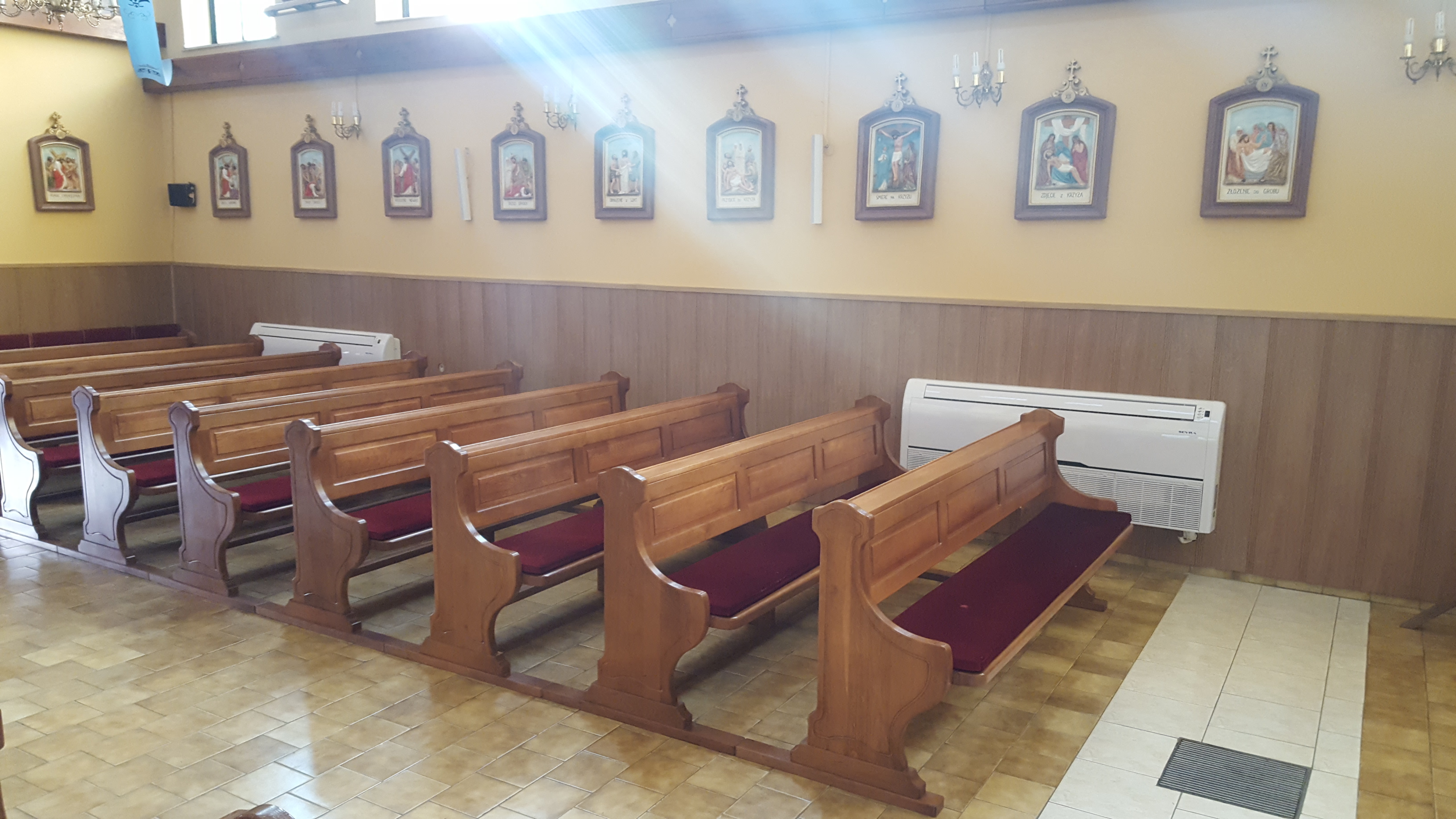 Wnętrze kościoła z drewnianymi ławkami i zamontowaną jednostką klimatyzacyjną na ścianie, oświetloną światłem słonecznym wpadającym przez okno.