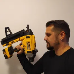 Mężczyzna z brodą prezentuje żółto-czarną, akumulatorową gwoździarkę marki DeWalt, trzymając ją w dłoni na tle jasnej ściany.