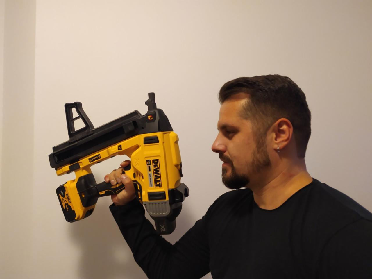 Mężczyzna z brodą prezentuje żółto-czarną, akumulatorową gwoździarkę marki DeWalt, trzymając ją w dłoni na tle jasnej ściany.