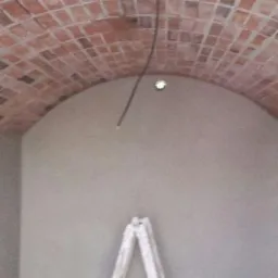 Wnętrze pomieszczenia z łukowym sufitem wykończonym cegłą, ściany świeżo pokryte gładzią, drabina oparta o ścianę, widoczny przewód instalacji elektrycznej wystający z sufitu.