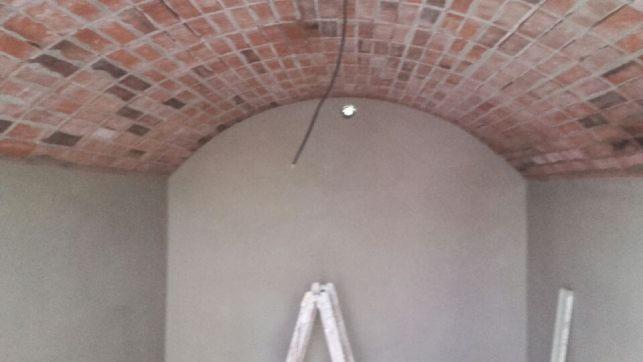 Wnętrze pomieszczenia z łukowym sufitem wykończonym cegłą, ściany świeżo pokryte gładzią, drabina oparta o ścianę, widoczny przewód instalacji elektrycznej wystający z sufitu.