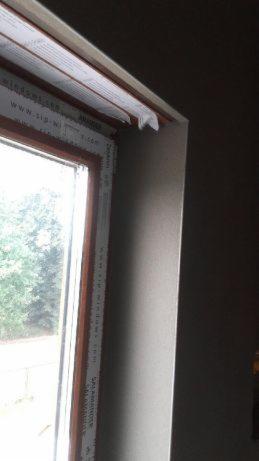 Fragment ściany z oknem po nałożeniu gładzi, widoczne zabezpieczenia okna folią i taśmą malarską.