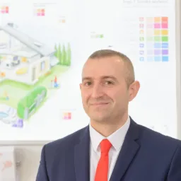 Uśmiechnięty broker ubezpieczeniowy w garniturze, na tle tablicy z planem domu i paletą kolorów. Prezentacja ubezpieczenia domu i życia.