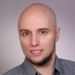 Portret mężczyzny z krótką brodą, ubrany w koszulę w kratę na szarym tle. Uśmiech na twarzy, jasne oczy, oświetlenie studyjne.