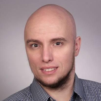 Portret mężczyzny z krótką brodą, ubrany w koszulę w kratę na szarym tle. Uśmiech na twarzy, jasne oczy, oświetlenie studyjne.