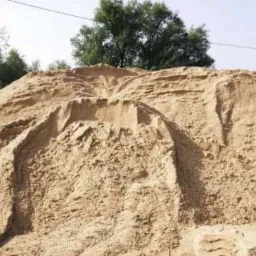 Duża pryzma piasku budowlanego na tle drzew i nieba, widoczne ślady po zsypywaniu materiału.