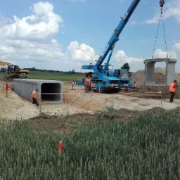 Montaż prefabrykowanych elementów żelbetowych w Brusach: dźwig podnosi betonowy moduł o kwadratowym przekroju, w tle wykopany rów z ułożonym już fragmentem konstrukcji, widoczny również żółty koparka.