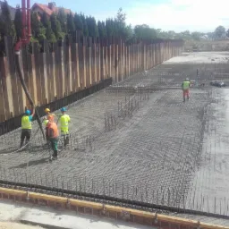 Wylewanie betonu na zbrojenie fundamentu płyty w otoczeniu ścianki berlińskiej, widoczni pracownicy w odblaskowych kamizelkach i kaskach.