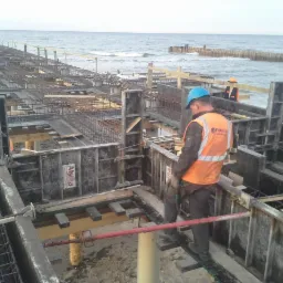 Prace zbrojarskie i betonowe przy budowie konstrukcji żelbetowej na brzegu morza, widoczne szalunki firmy Alsina i zbrojenia, pracownik w kasku i kamizelce odblaskowej.
