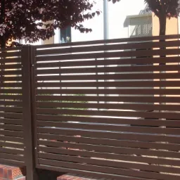 Nowoczesne ogrodzenie z poziomych, brązowych paneli metalowych, z widocznym fragmentem murku z czerwonej cegły i drzewem w tle.
