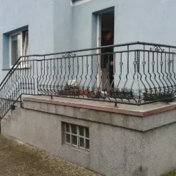 Czarna, metalowa balustrada z ozdobnymi elementami na schodach i balkonie jasnoniebieskiego domu. W tle widoczna osoba w drzwiach.