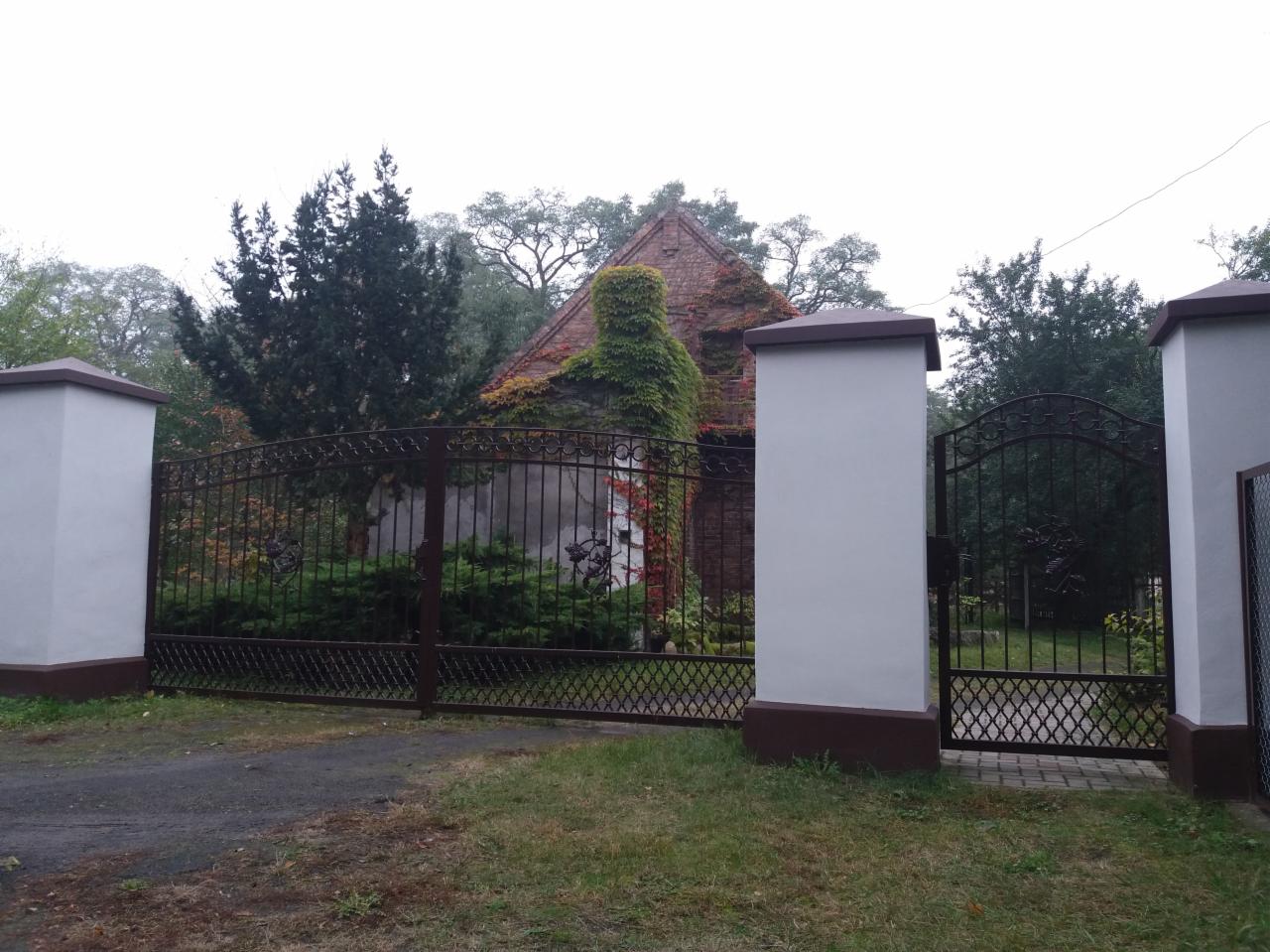 Kuta brama wjazdowa z dekoracyjnymi elementami, umieszczona między murowanymi słupkami z ciemnobrązowymi podstawami i nakryciami, prowadząca do zarośniętego bluszczem domu.