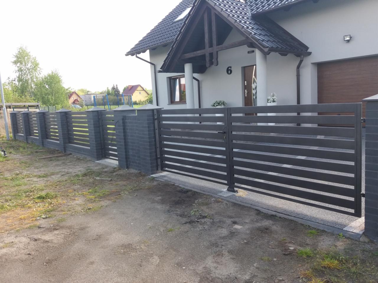 Nowoczesne ogrodzenie panelowe z poziomymi przęsłami i murowanymi słupkami w kolorze grafitowym, z bramą wjazdową, na tle domu z ciemną dachówką.