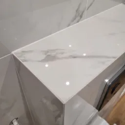 Wykończona półka łazienkowa z płytek imitujących marmur, z widocznym przyłączem wodnym i fragmentem zabudowy WC, fuga w kolorze białym.