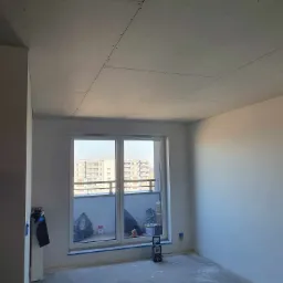 Pokój w trakcie remontu: surowy sufit z płyt kartonowo-gipsowych, ściany przygotowane do malowania, okno balkonowe z widokiem na osiedle, widoczne ślady po pracach budowlanych na podłodze.