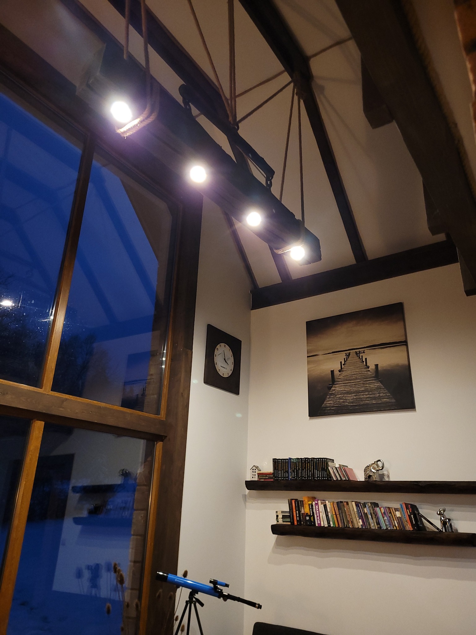 Wnętrze pokoju z dużym oknem, drewnianymi belkami sufitowymi i oświetleniem punktowym, z półkami na książki i obrazem mola na ścianie.