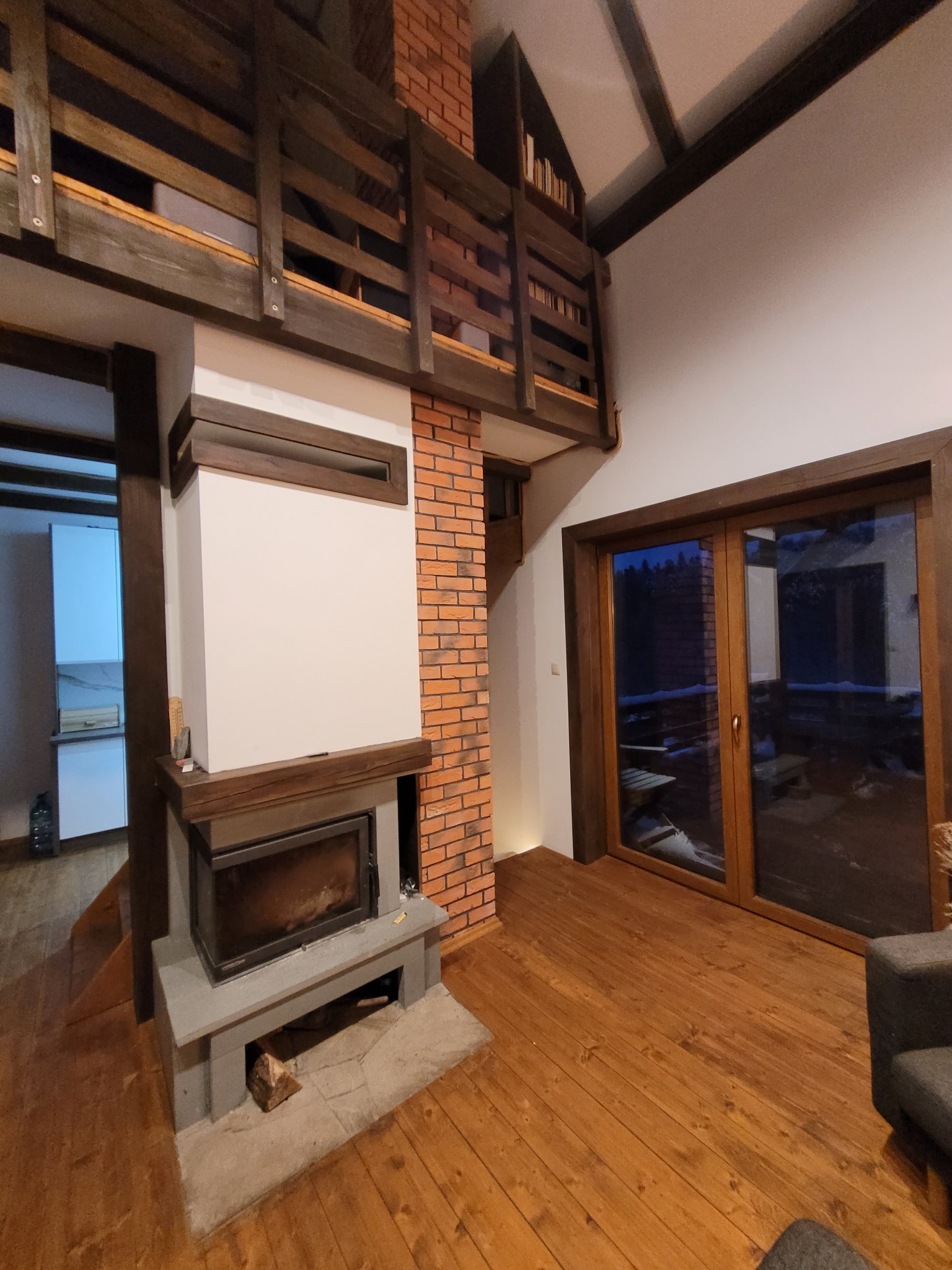 Wnętrze salonu z kominkiem obudowanym cegłą, drewnianą podłogą i antresolą z drewnianą balustradą, widok na podwójne drzwi balkonowe i częściowo widoczną kuchnię w tle.