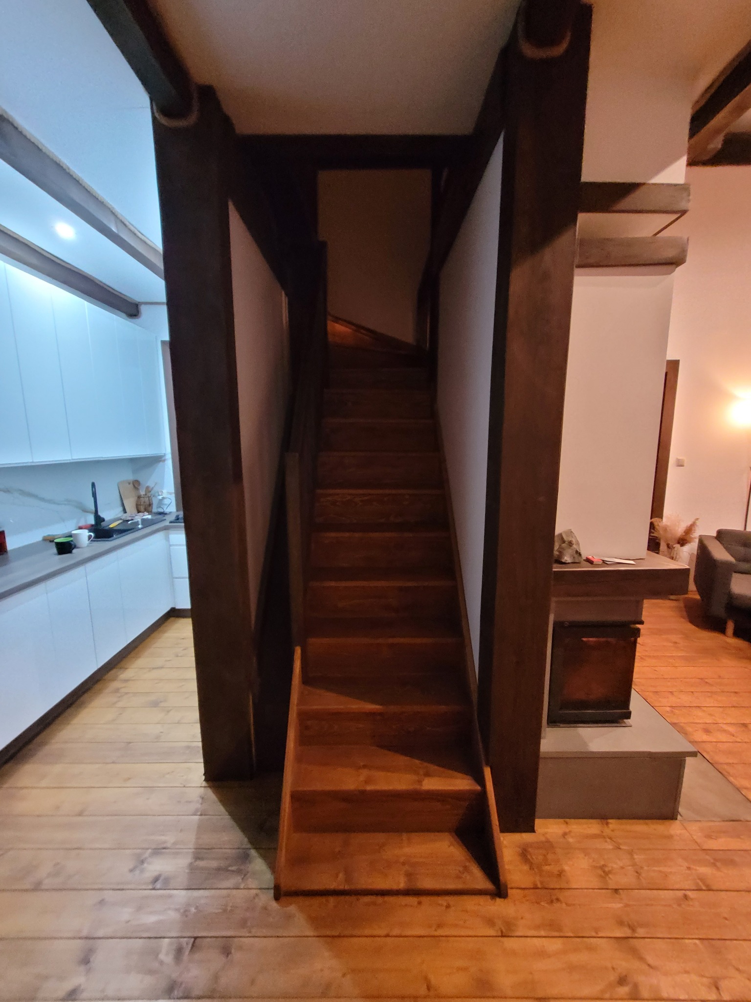 Wnętrze domu z drewnianymi schodami, widok na kuchnię z białymi szafkami i salon z kominkiem w tle.