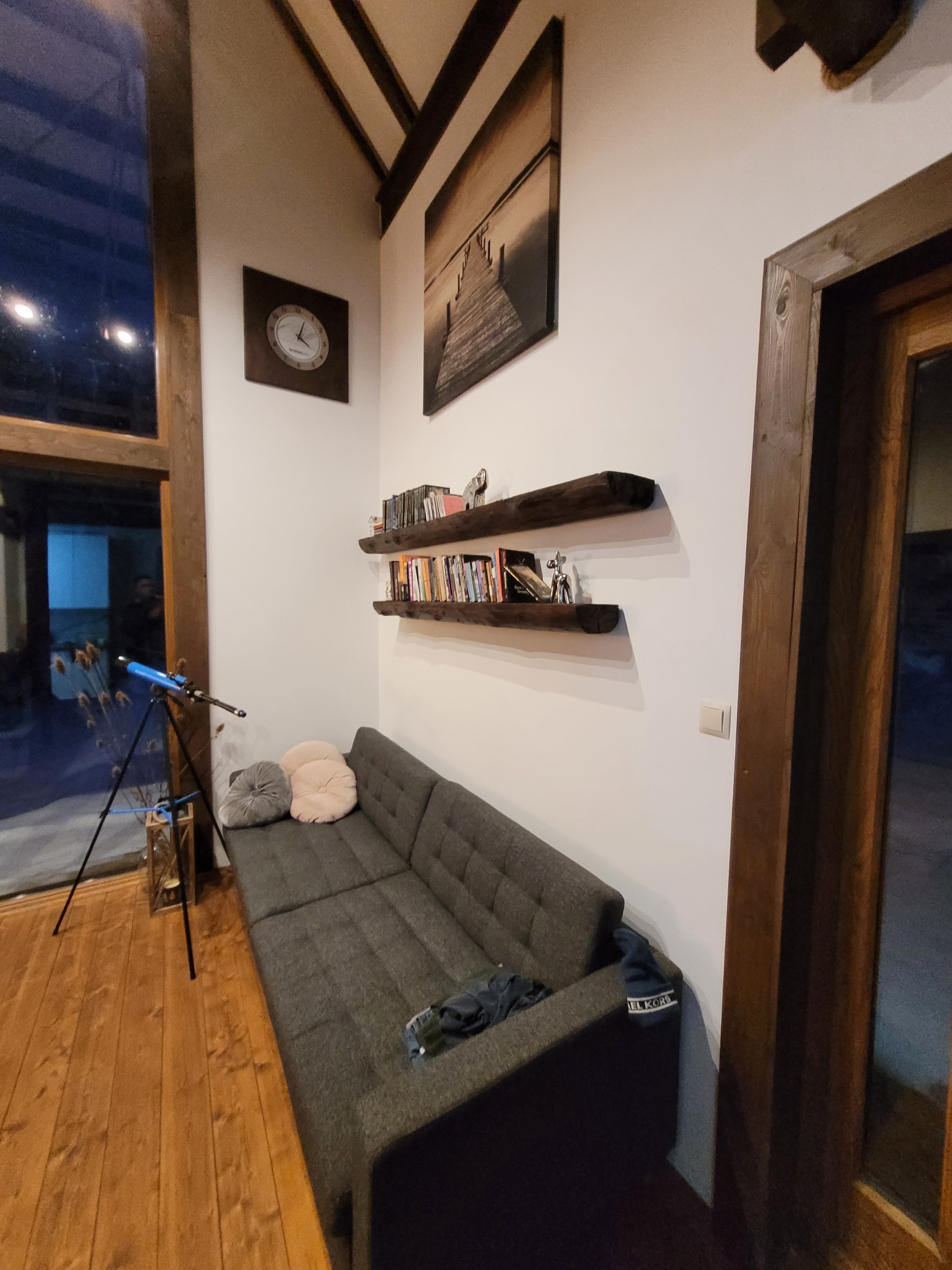Wnętrze pokoju z szarą sofą, drewnianymi półkami z książkami i dekoracjami, zegarem na ścianie oraz drewnianą stolarką okienną i drzwiową.
