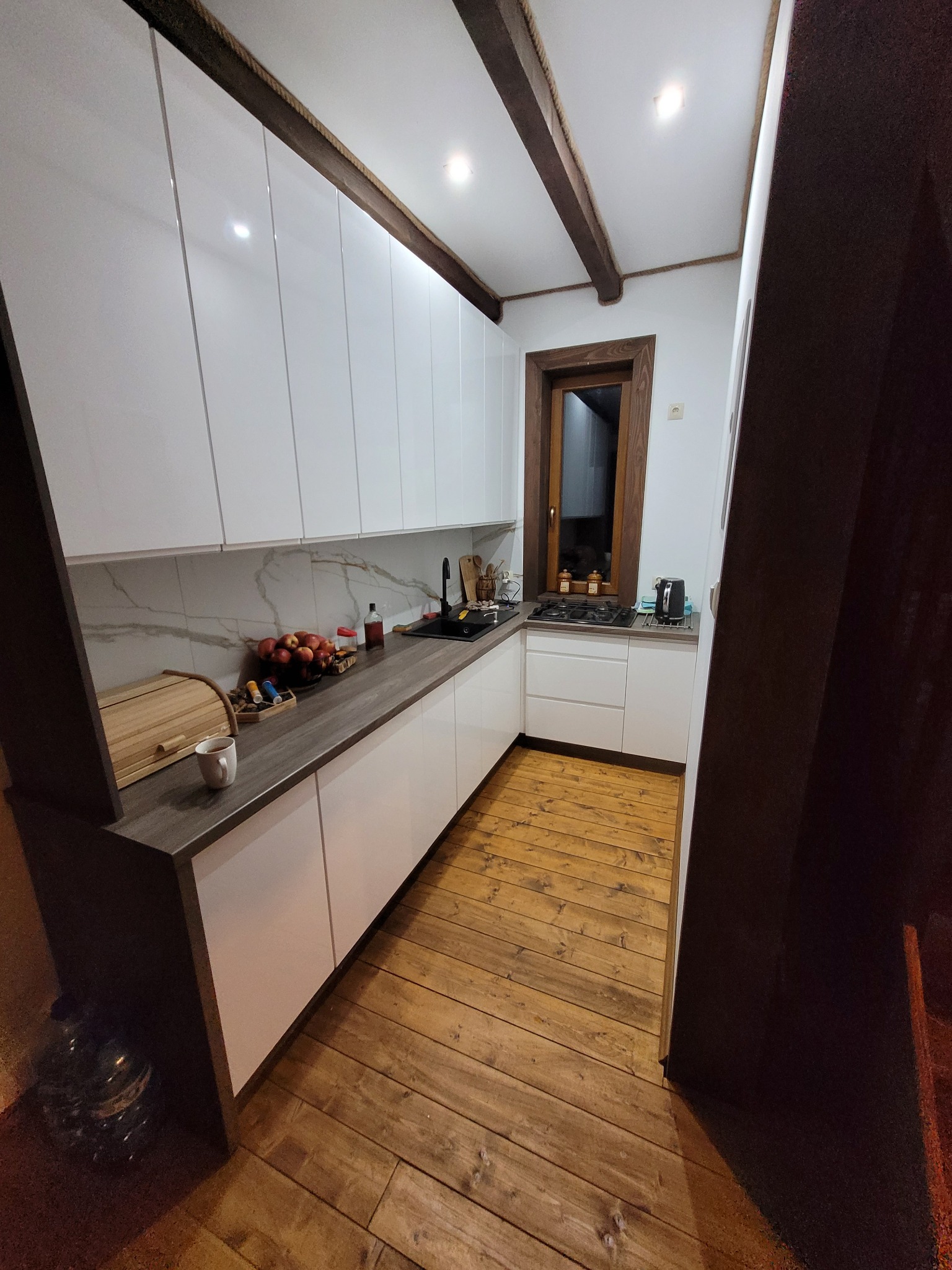 Nowoczesna kuchnia z białymi szafkami, drewnianym blatem, marmurowym backsplashiem i drewnianą podłogą, oświetlona punktowymi lampami sufitowymi i z widocznymi drewnianymi belkami stropowymi.