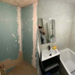 ALTIBUD - Łazienka w trakcie remontu: po lewej surowe ściany z resztkami tynku, po prawej wykończona ściana z jasnymi płytkami, umywalka z czarną armaturą i lustro z odbiciem osoby robiącej zdjęcie.