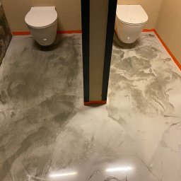 ALTIBUD - Biało-szara wylewka samopoziomująca w łazience z dwoma toaletami, zabezpieczona pomarańczową taśmą malarską przy ścianach i słupie konstrukcyjnym.