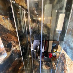 ALTIBUD - Nowoczesna kabina prysznicowa z transparentnymi szklanymi drzwiami i marmurowymi ścianami, widoczne odbicie osoby wykonującej zdjęcie, czarne wiadro i czerwona szmatka w rogu kabiny.