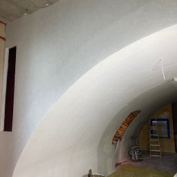 ALTIBUD - Wnętrze z łukowym sufitem po nałożeniu gładzi, widoczne wiaderka z farbą, drabina i zakryta folią podłoga.