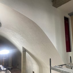 ALTIBUD - Wnętrze w trakcie prac wykończeniowych, widoczna gładź na łukowym suficie, rusztowanie i lampa budowlana, podłoga zabezpieczona folią.