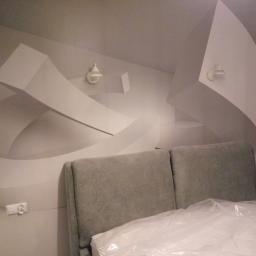 ALTIBUD - Nowoczesna sypialnia w odcieniach szarości z geometrycznym malowaniem ściennym, obejmującym abstrakcyjne formy i dwa kinkiety, z widocznym fragmentem łóżka z szarym zagłówkiem i materacem w folii.