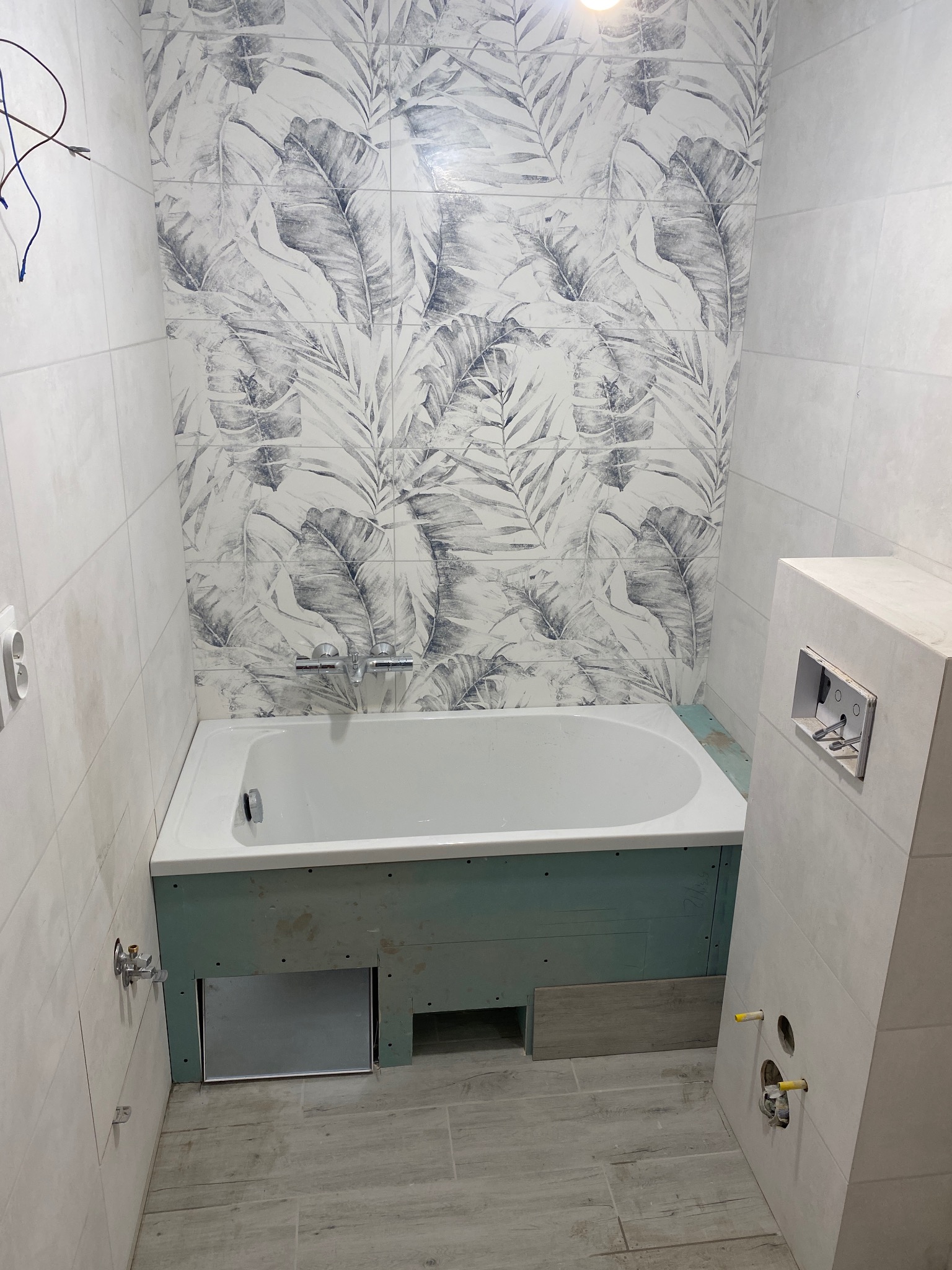 Łazienka w trakcie wykończenia: biała wanna obudowana płytą gipsowo-kartonową, ściany pokryte białymi płytkami z motywem liści, widoczne elementy instalacji wodnej i elektrycznej.