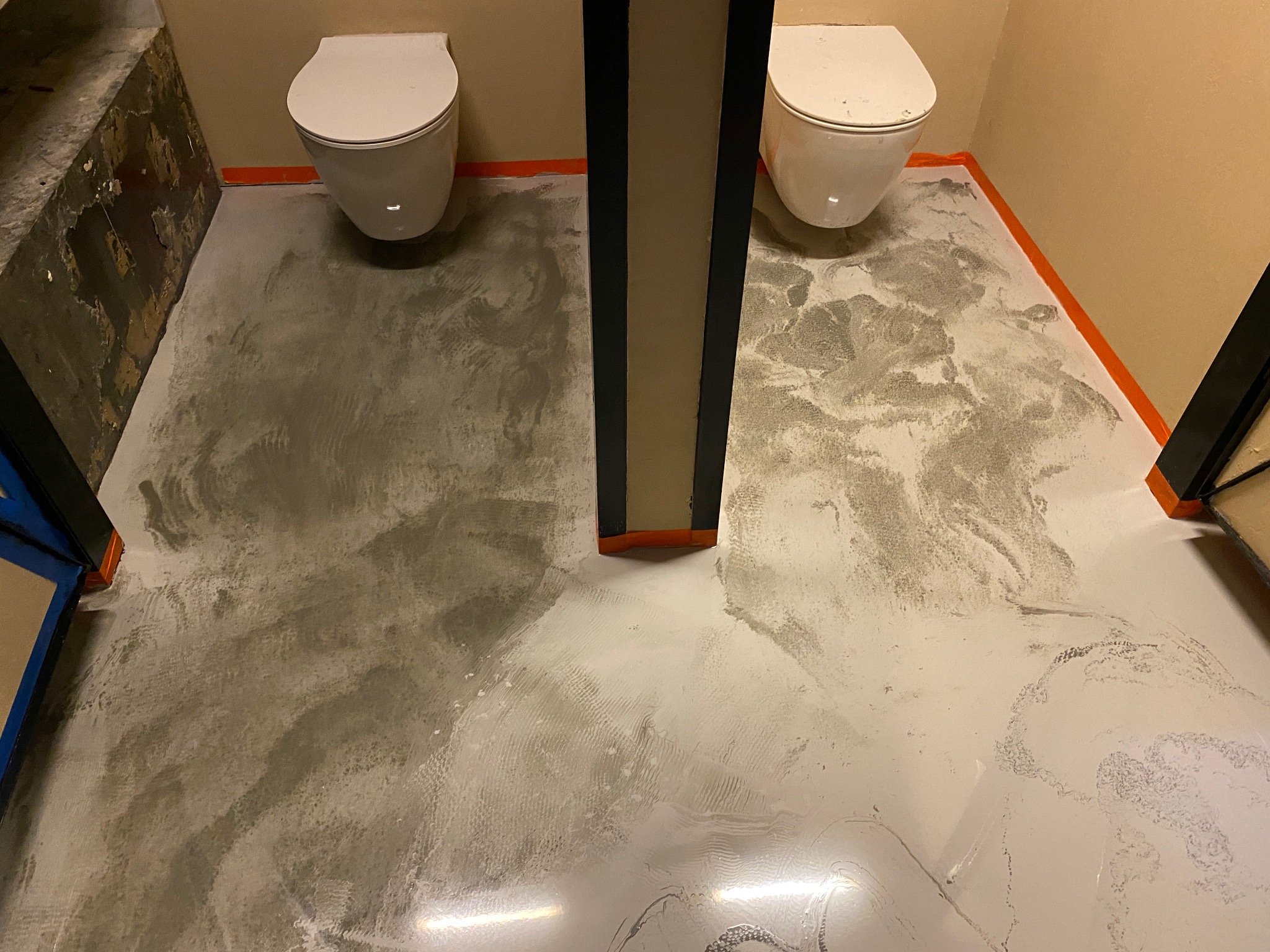 Biało-szara wylewka samopoziomująca w łazience z dwoma toaletami, zabezpieczona pomarańczową taśmą malarską przy ścianach i słupie konstrukcyjnym.