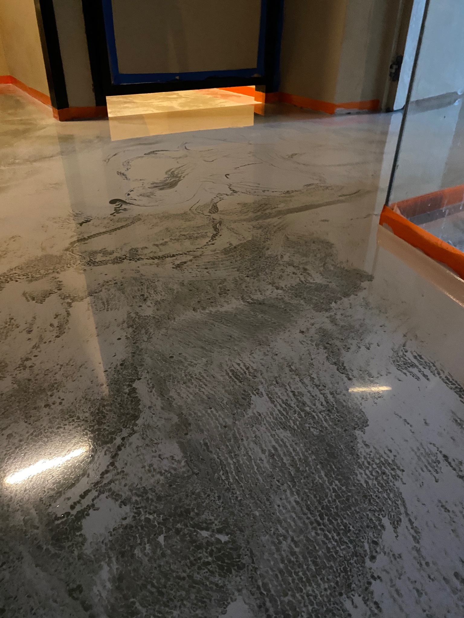 Świeżo wylana, połyskująca posadzka żywiczna z widocznymi smugami i wzorami, zabezpieczona taśmą malarską przy ścianach i drzwiach.
