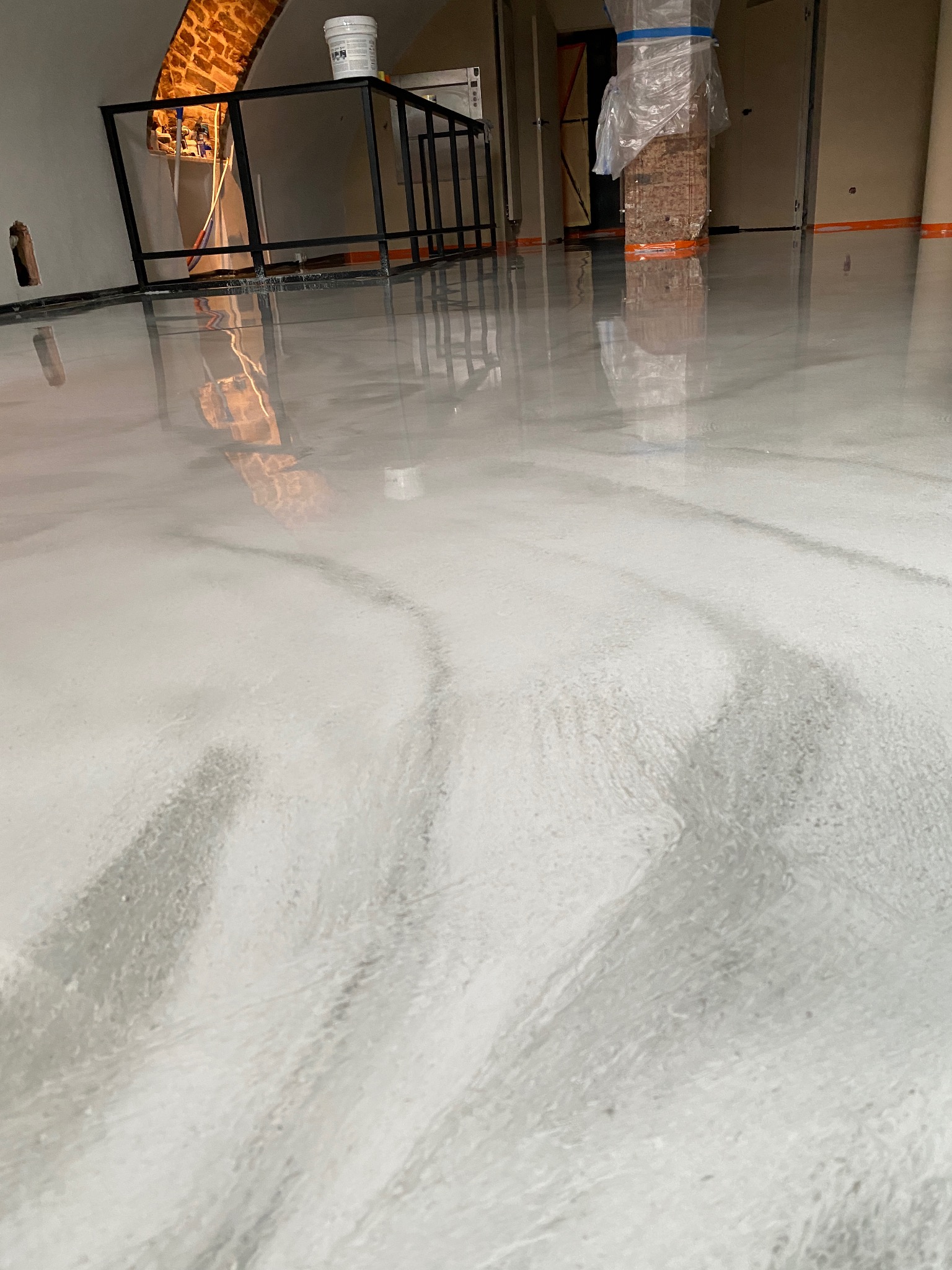 Świeżo wylana, samopoziomująca podłoga w odcieniach szarości z widocznymi smugami, odbijająca elementy wnętrza: metalową konstrukcję, fragment ceglanego łuku i filar zabezpieczony folią. W tle...