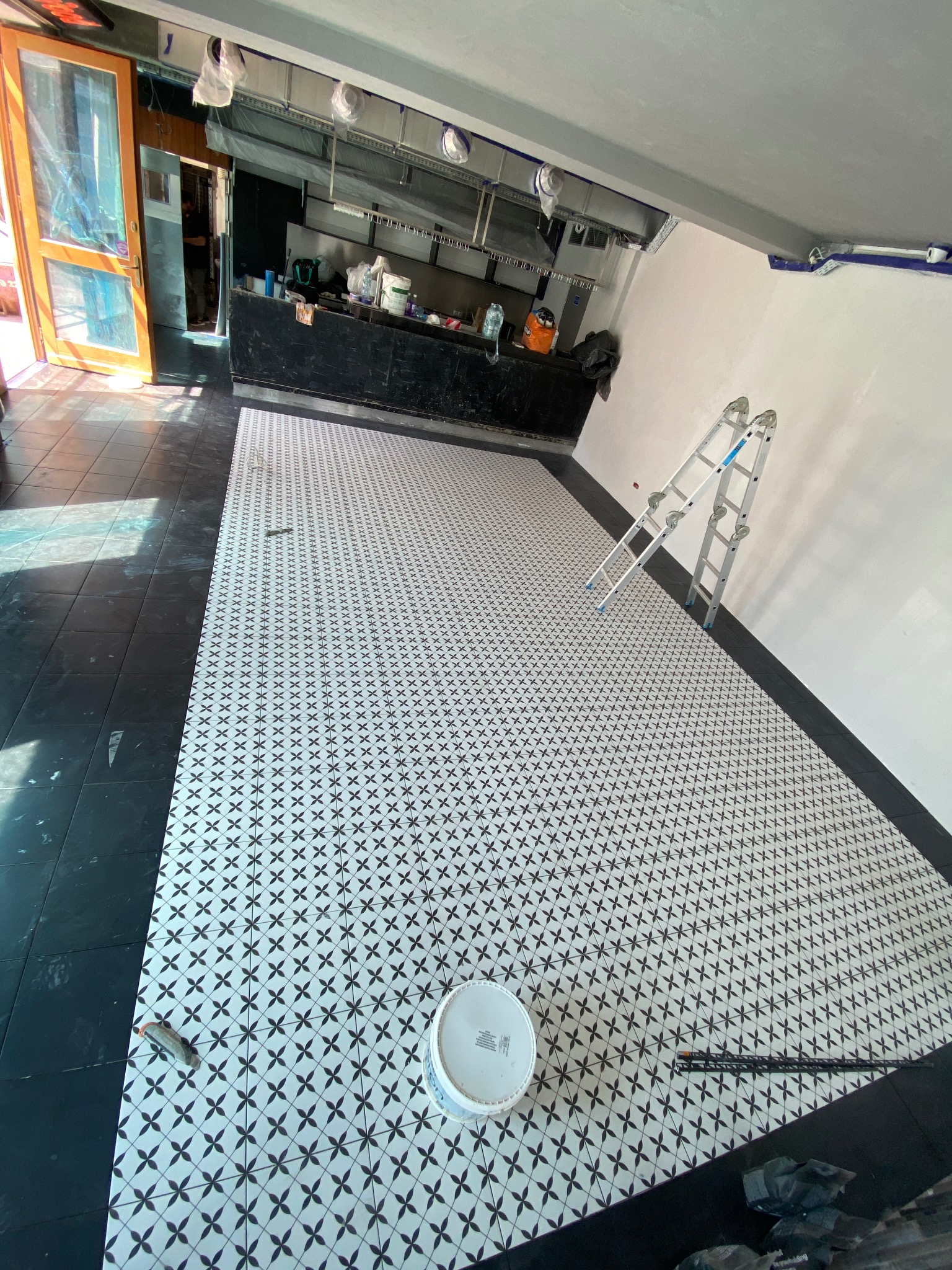 Układanie białych płytek podłogowych z czarnym wzorem w lokalu użytkowym, widoczna drabina aluminiowa oparta o ścianę, wiadro z zaprawą i narzędzia glazurnicze.