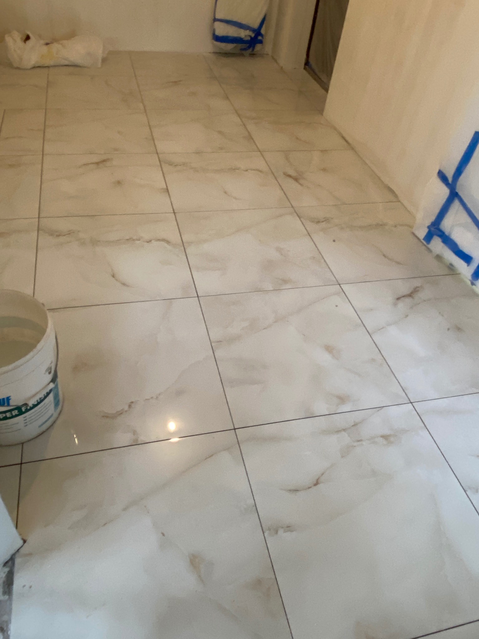 Ułożone, jasne płytki podłogowe imitujące marmur z widocznym wiadrem farby w rogu pomieszczenia i zabezpieczonymi taśmą malarską ścianami.