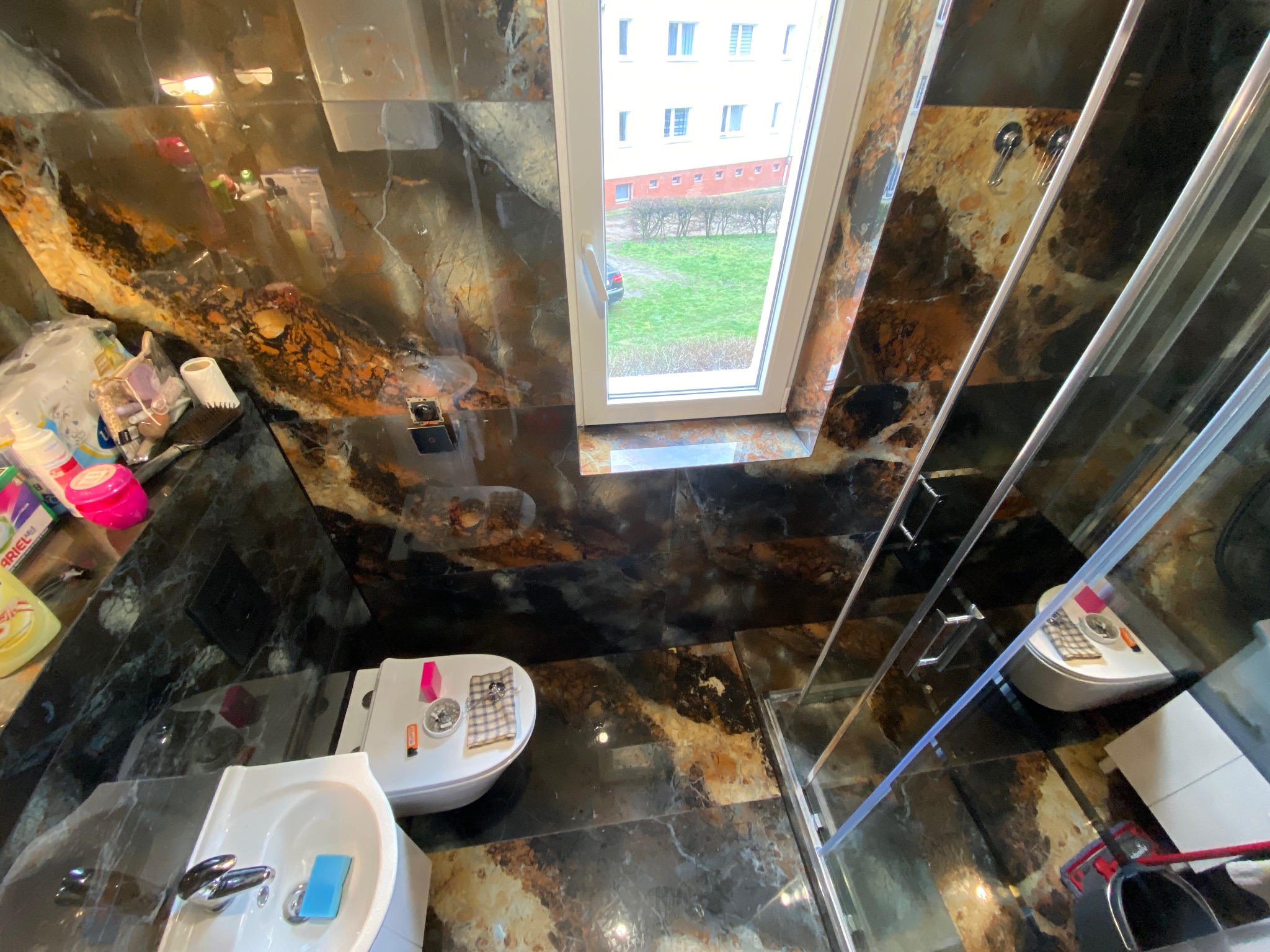 Wnętrze łazienki z marmurowymi płytkami na ścianach i podłodze, widok z góry na umywalkę, toaletę i kabinę prysznicową z widokiem na okno