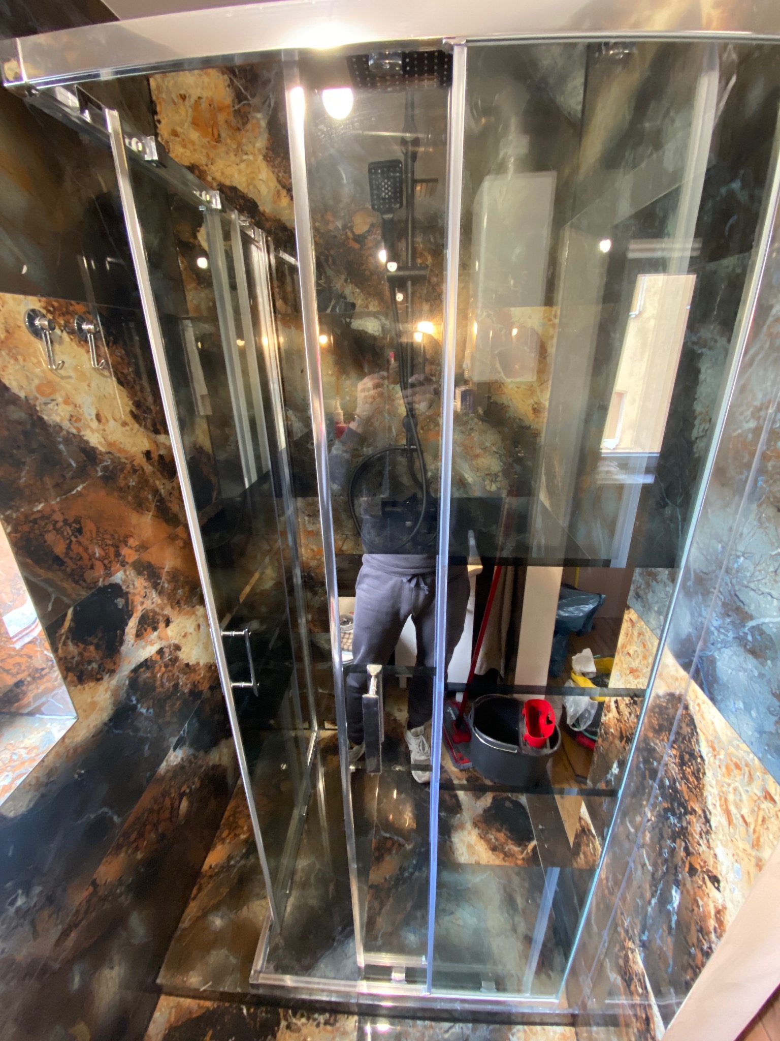 Nowoczesna kabina prysznicowa z transparentnymi szklanymi drzwiami i marmurowymi ścianami, widoczne odbicie osoby wykonującej zdjęcie, czarne wiadro i czerwona szmatka w rogu kabiny.