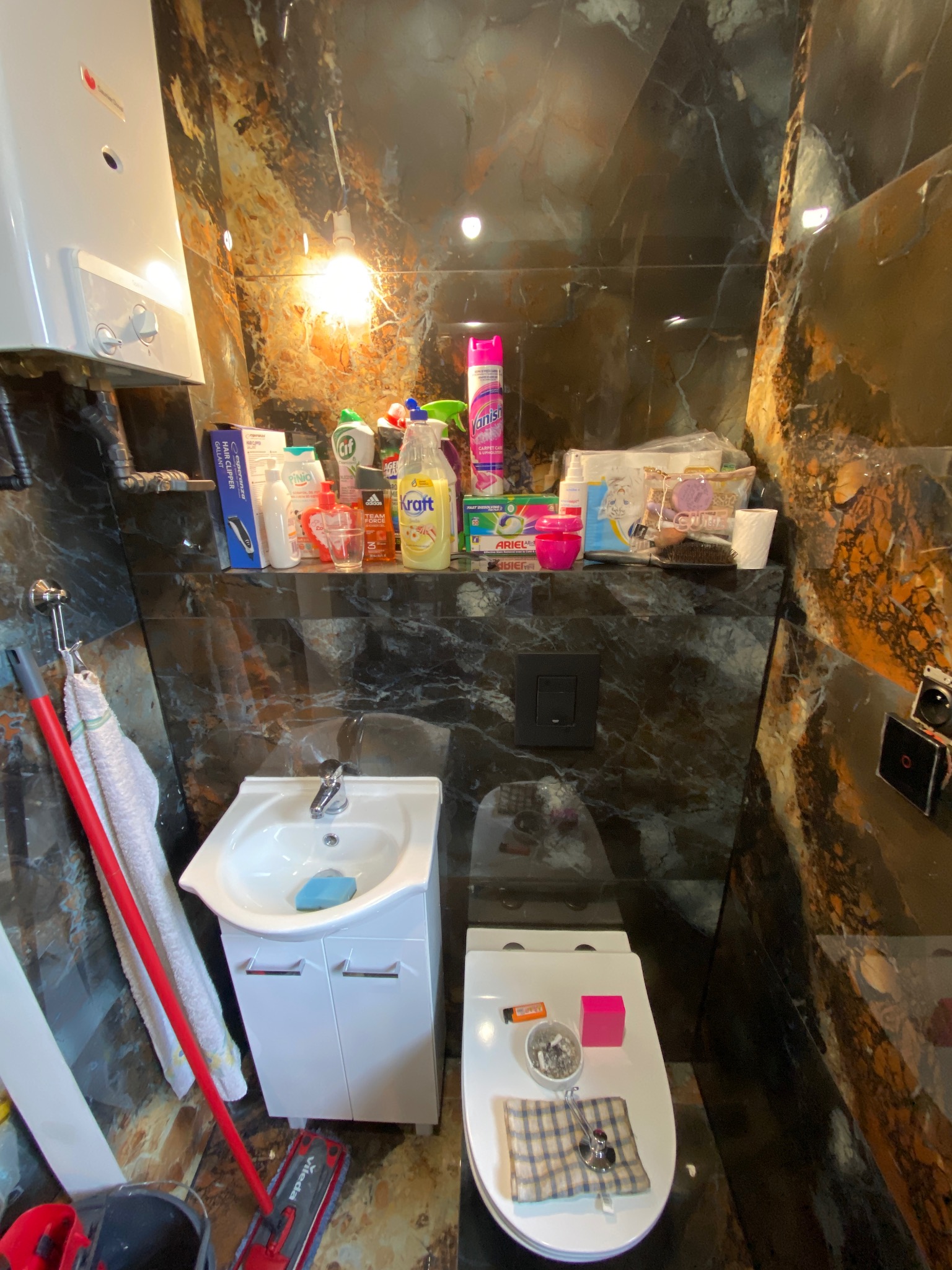 Wnętrze łazienki z umywalką, toaletą i półką z różnymi środkami czystości oraz akcesoriami na tle marmurowych ścian. Na toalecie leży szmatka w kratę i popielniczka z niedopałkami.