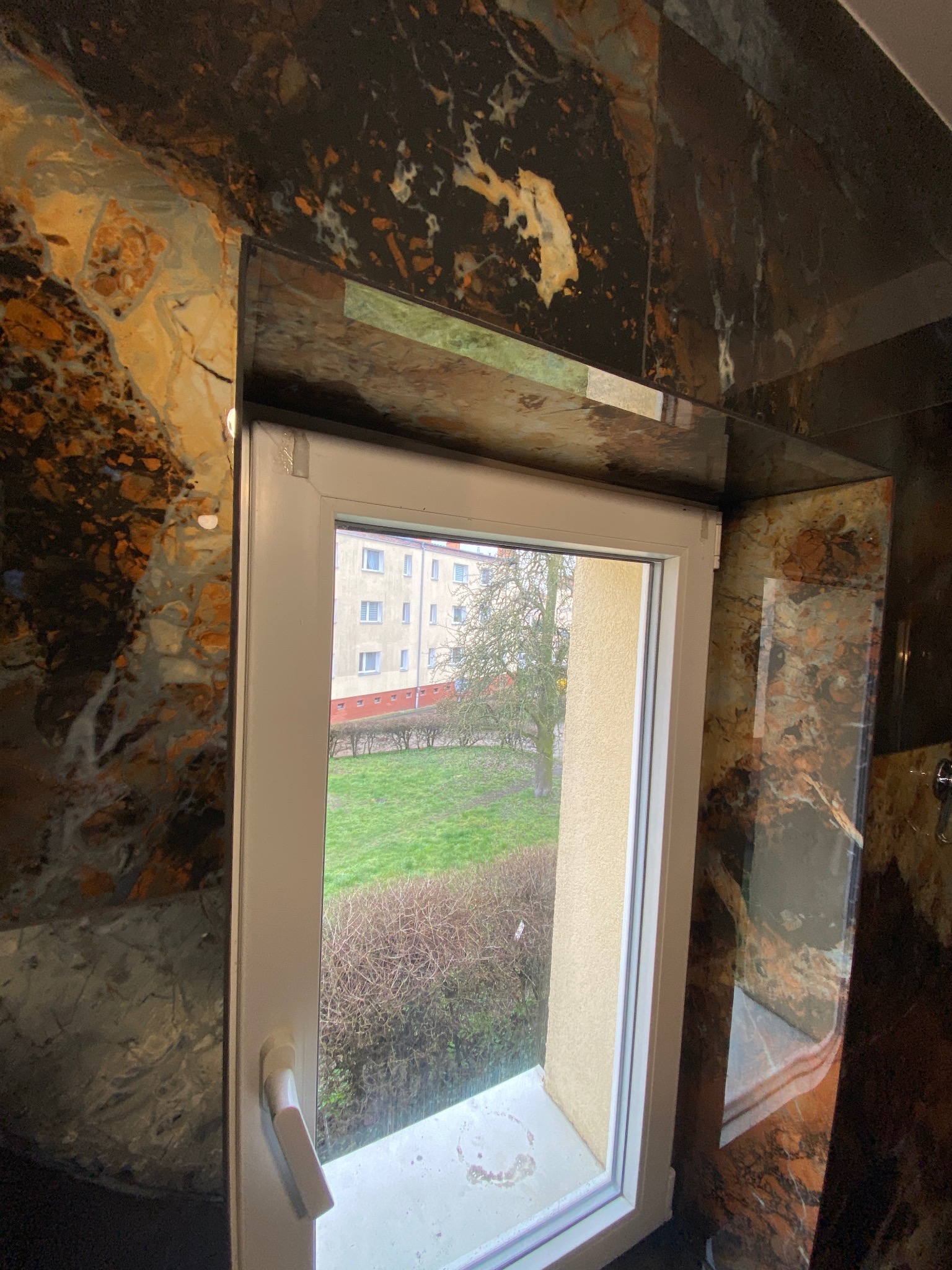 Okno PCV z widokiem na zieleń, osadzone w ścianie wykończonej płytami imitującymi ciemny marmur z jasnymi żyłami.